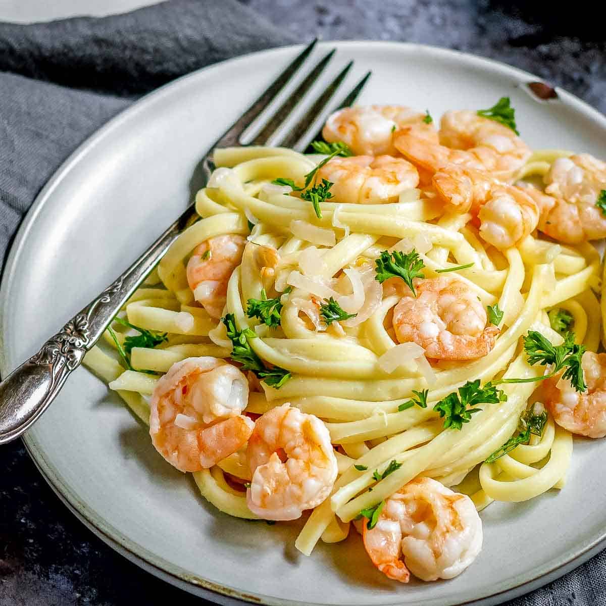 Side close up shot of shrimp scampi garnished with parsley on a grey plate with a fork on a black and grey surface.