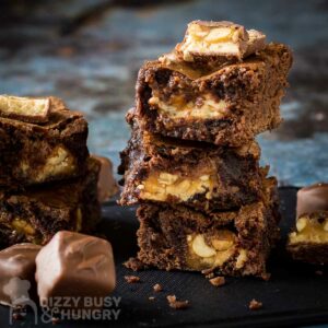 Side shot of multiple snickers brownies stacked on each other with sliced snickers bars on top with full snickers bars on the side all on a black surface.
