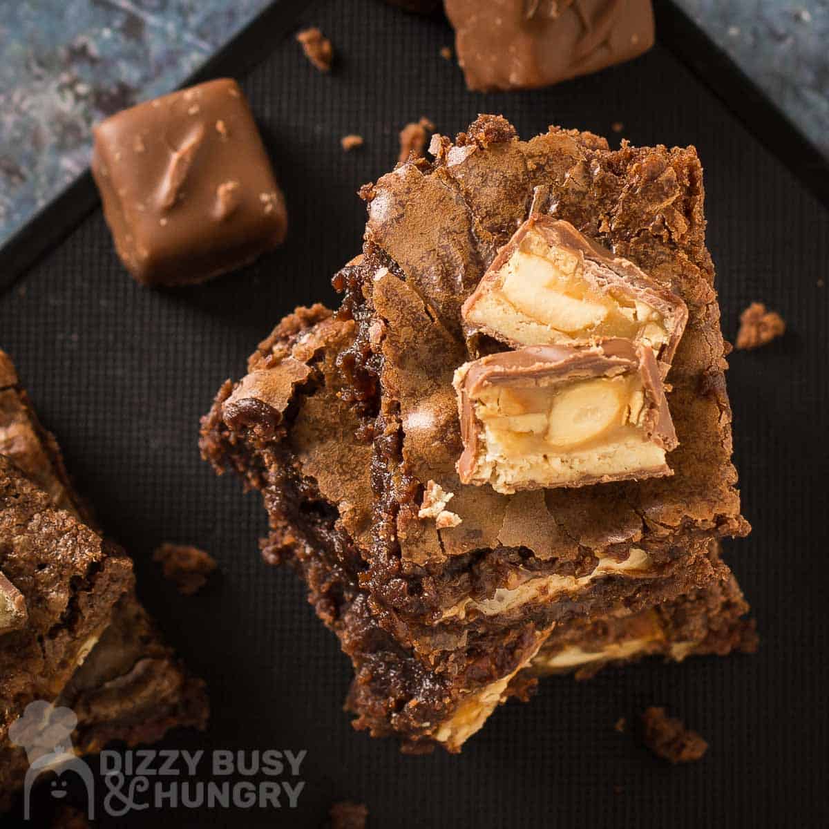 Overhead shot of three snickers brownies stacked on each other with sliced snickers bars on top with full snickers bars on the side all on a black surface.