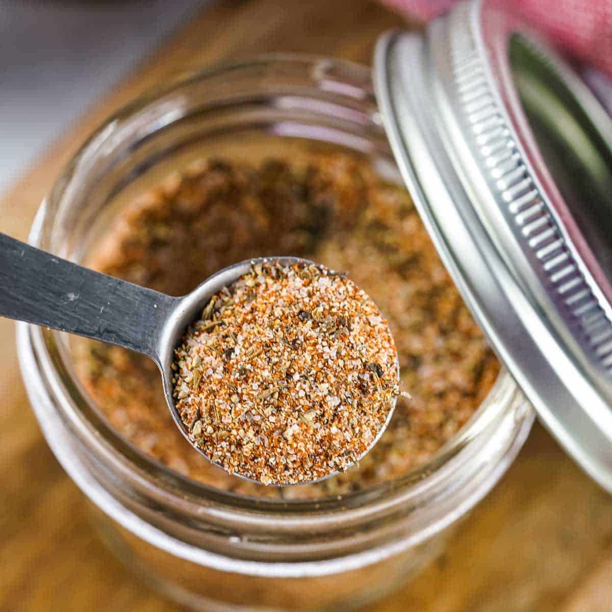 Close up overhead shot of a tablespoon holding some steak rub over a clear mason jar full of more steak rub with the lid leaning on the side.
