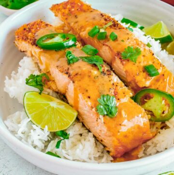 Side close up shot of two pieces of salmon on a bed of rice garnished with parsley and cut jalapeños in a large white bowl with lime wedges on the side.