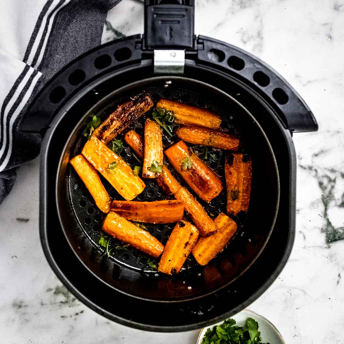 Overhead shot of roasted carrots with herbs in the air fryer on a marble countertop with a black and white striped cloth on the side.