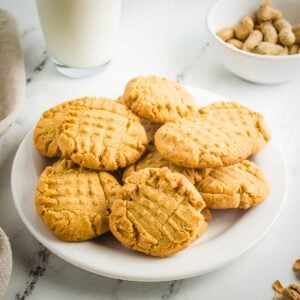 Side shot of multiple peanut butter cookies arranged on a white plate on a white marble surface with a glass of milk and a white bowl with whole peanuts in the background.