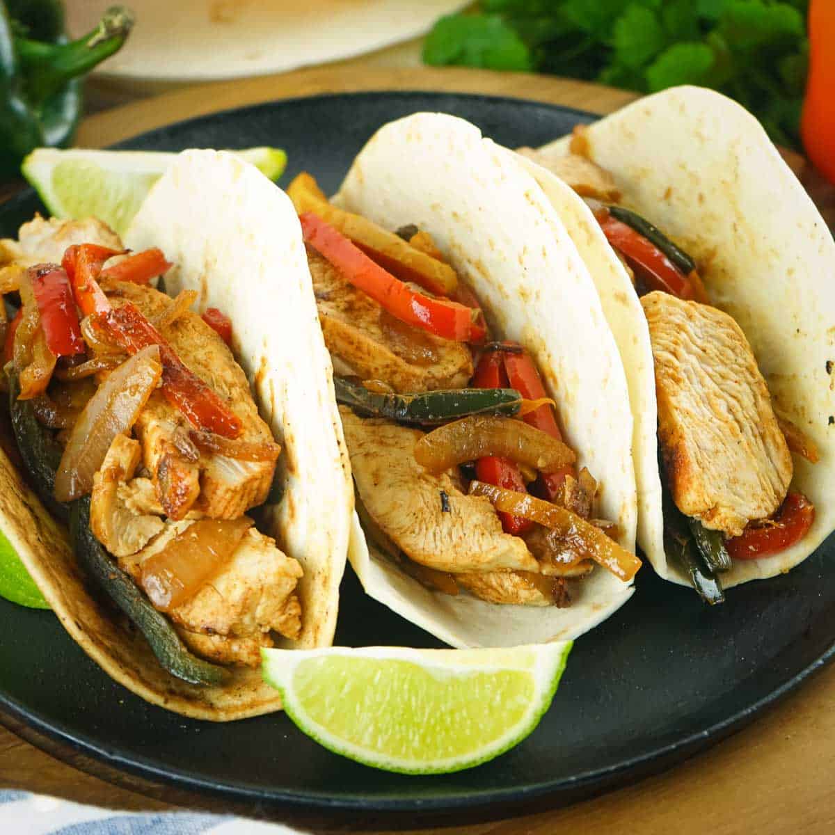 Side shot of three chicken fajita tacos on a black plate with a lime wedge on the side and fresh herbs in the background.