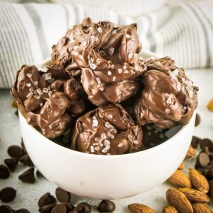 Side shot of multiple chocolate nut clusters with sea salt in a white bowl with almonds and chocolate chips sprinkled around.
