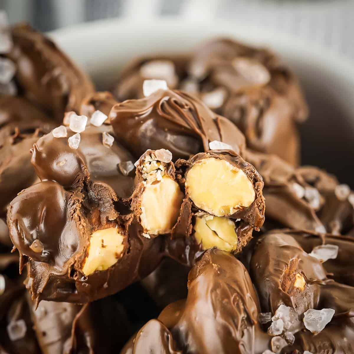 Close up side shot of one chocolate nut cluster with a bite taken out in a white bowl with other clusters sprinkled with sea salt.