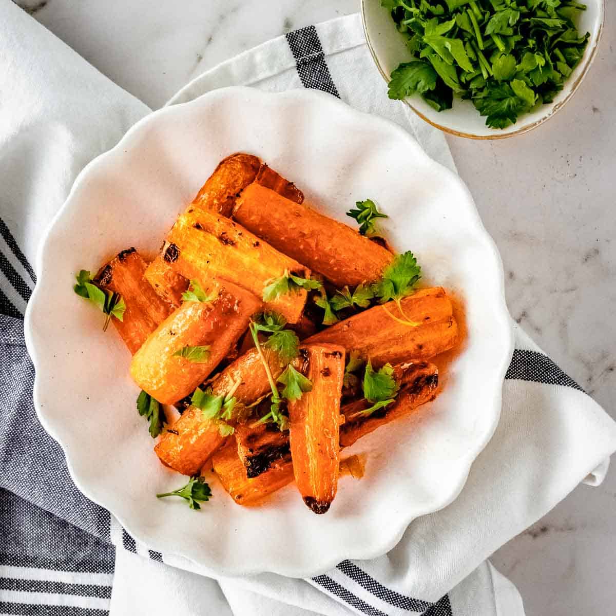 Overhead shot of roasted carrots garnished with herbs on a white plate on a grey and white cloth with a small bowl of herbs on the side.