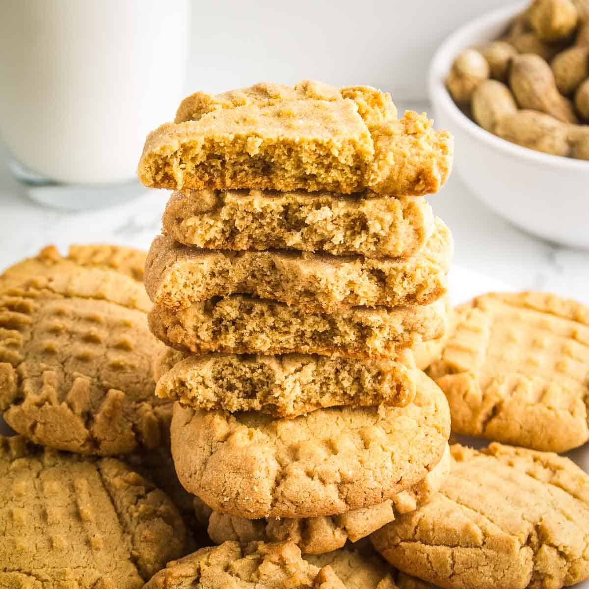 Side shot of multiple halved peanut butter cookies stacked on each other on a white marble surface with a glass of milk and a white bowl with whole peanuts in the background.
