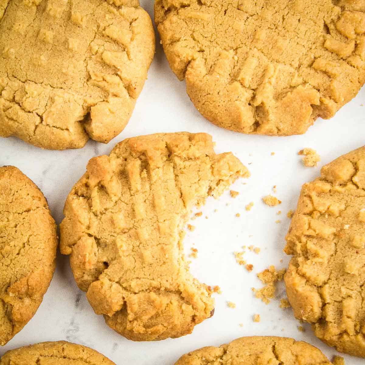 Overhead close up shot of multiple peanut butter cookies, one with a bite taken out, on a white marble surface.