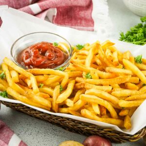 Side shot of French fries garnished with herbs in a basket lined with parchment paper and ketchup on the side in a clear bowl.