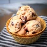 Side shot of multiple scoops of ice cream with cranberries in a double layered waffle cup sitting on a black and white striped cloth.