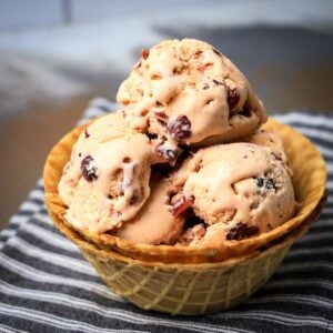 Side shot of multiple scoops of ice cream with cranberries in a double layered waffle cup sitting on a black and white striped cloth.