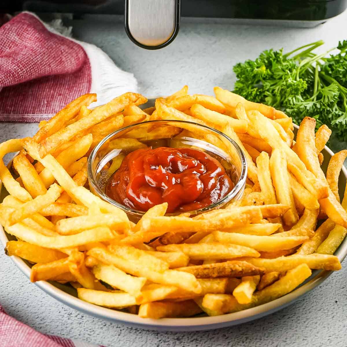 Side shot of golden French fries in a circle around a small clear bowl of ketchup with herbs and the air fryer in the background.