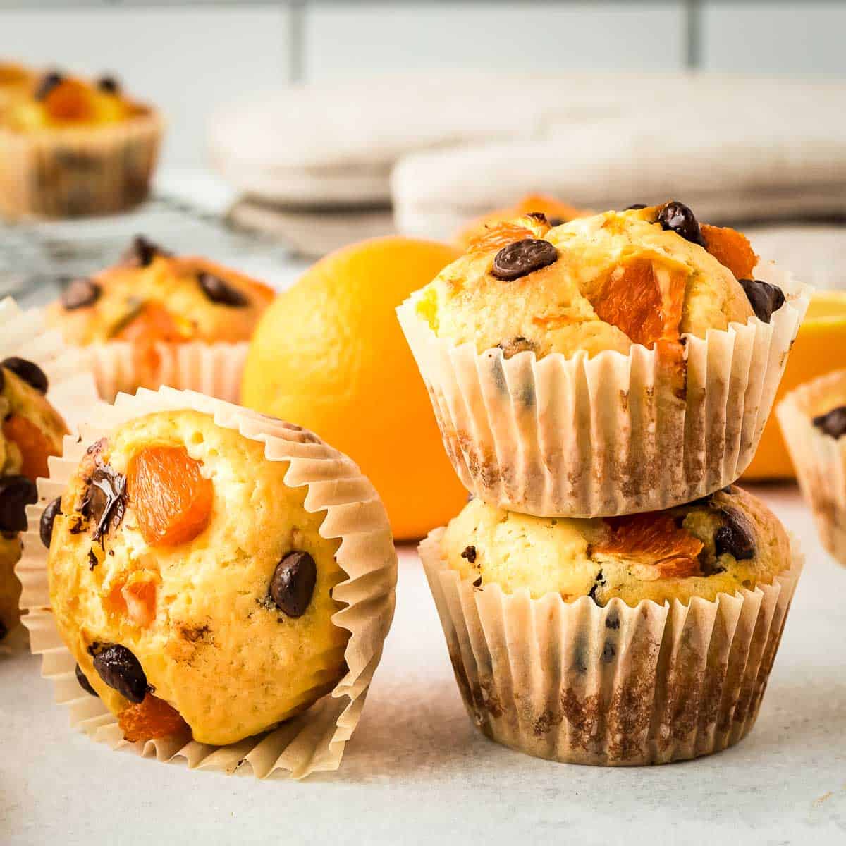 Side view of two stacked muffins and one turned on its side on a white surface with an orange and more muffins in the background.