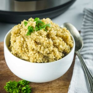 Side shot of a white bowl of fluffy quinoa garnished with fresh herbs on a wooden surface with a spoon and cloth napkin on the side.