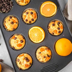 Overhead shot of a cupcake sheet filled with orange muffins, half slices of oranges, and chocolate chips.