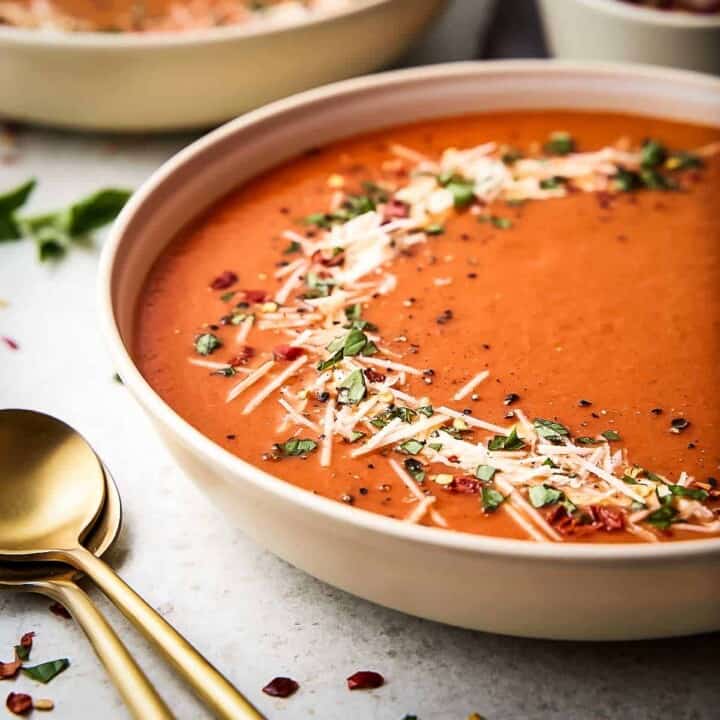 Side shot of a large white bowl with red pepper soup garnished with fresh herbs and parmesan cheese with gold spoons on the side.