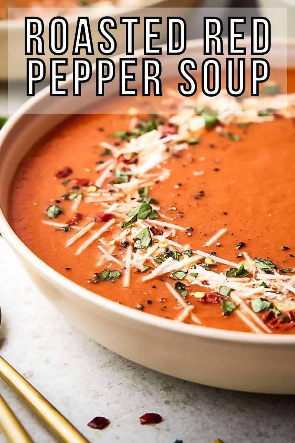 Side shot of a large white bowl with red pepper soup garnished with fresh herbs and parmesan cheese with gold spoons on the side.