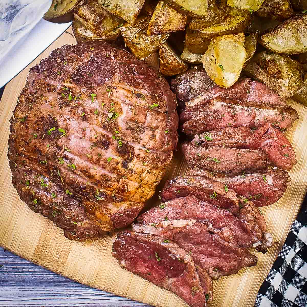 Overhead shot of a full lamb roast with slices of lamb and potato wedges on the side on a wooden cutting board.