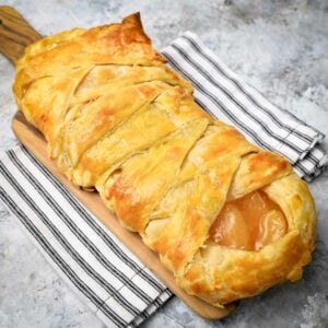 Side shot of criss-crossed apple danish on a small cutting board on a black and white striped cloth.