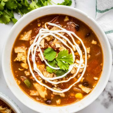Overhead shot of chicken tortilla soup in a white bowl garnished with swirls of sour cream and fresh herbs.