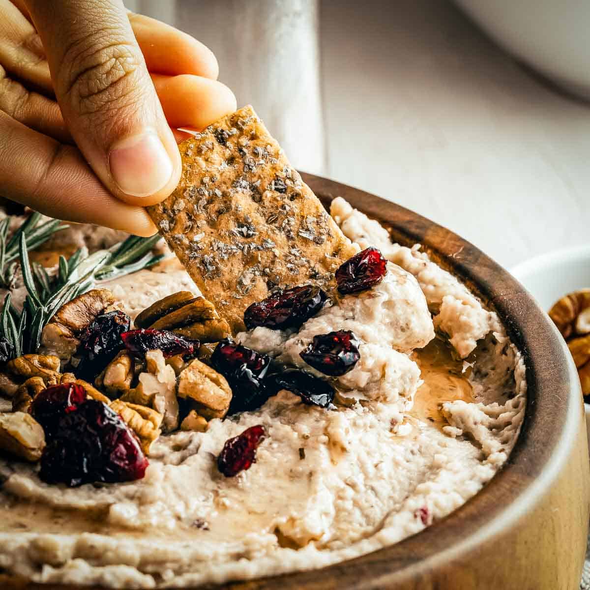 Side close up of a hand scooping some hummus with a cracker - the hummus is garnished with rosemary, cranberries, and pecans.