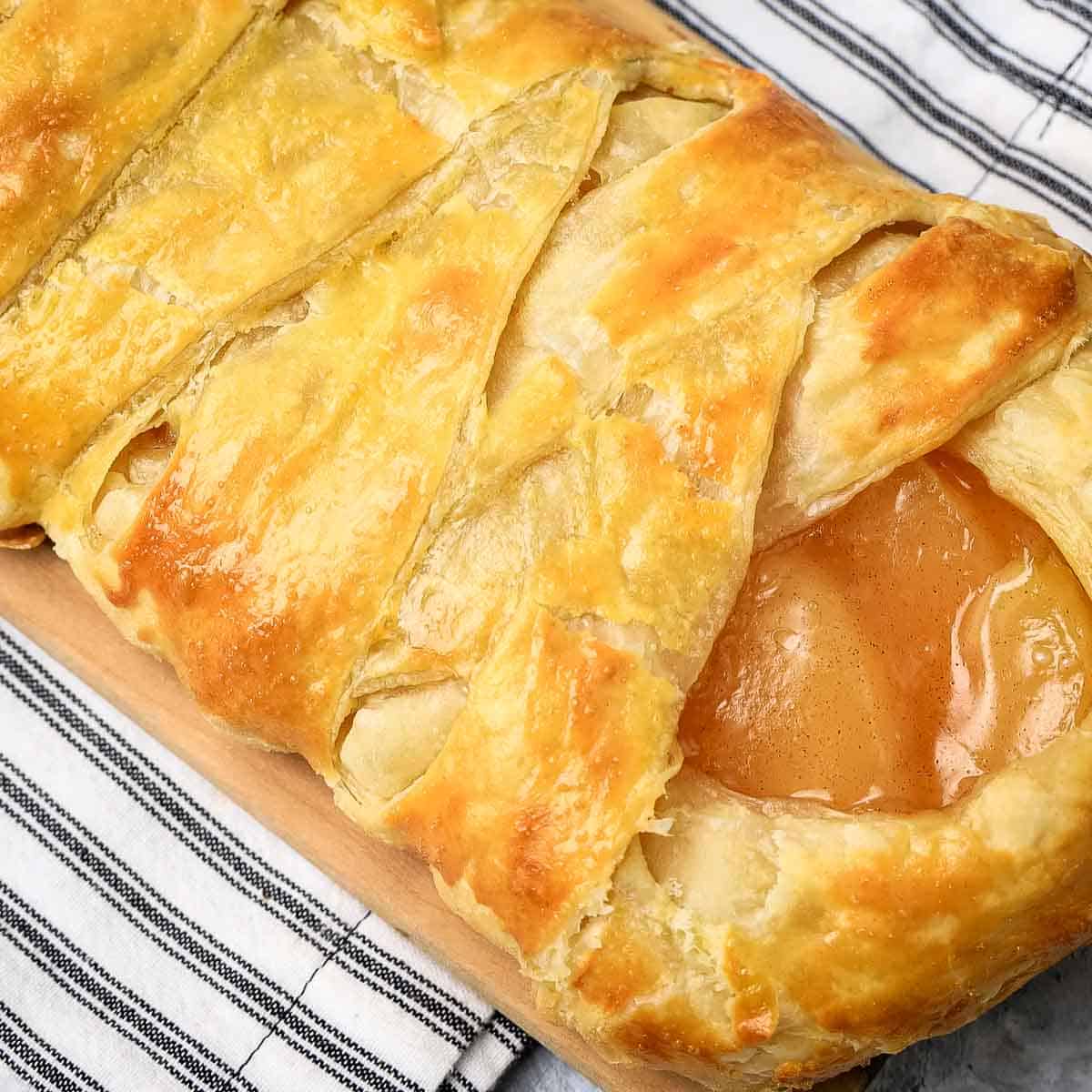 Side close up shot of criss-crossed apple danish on a small cutting board on a black and white striped cloth.