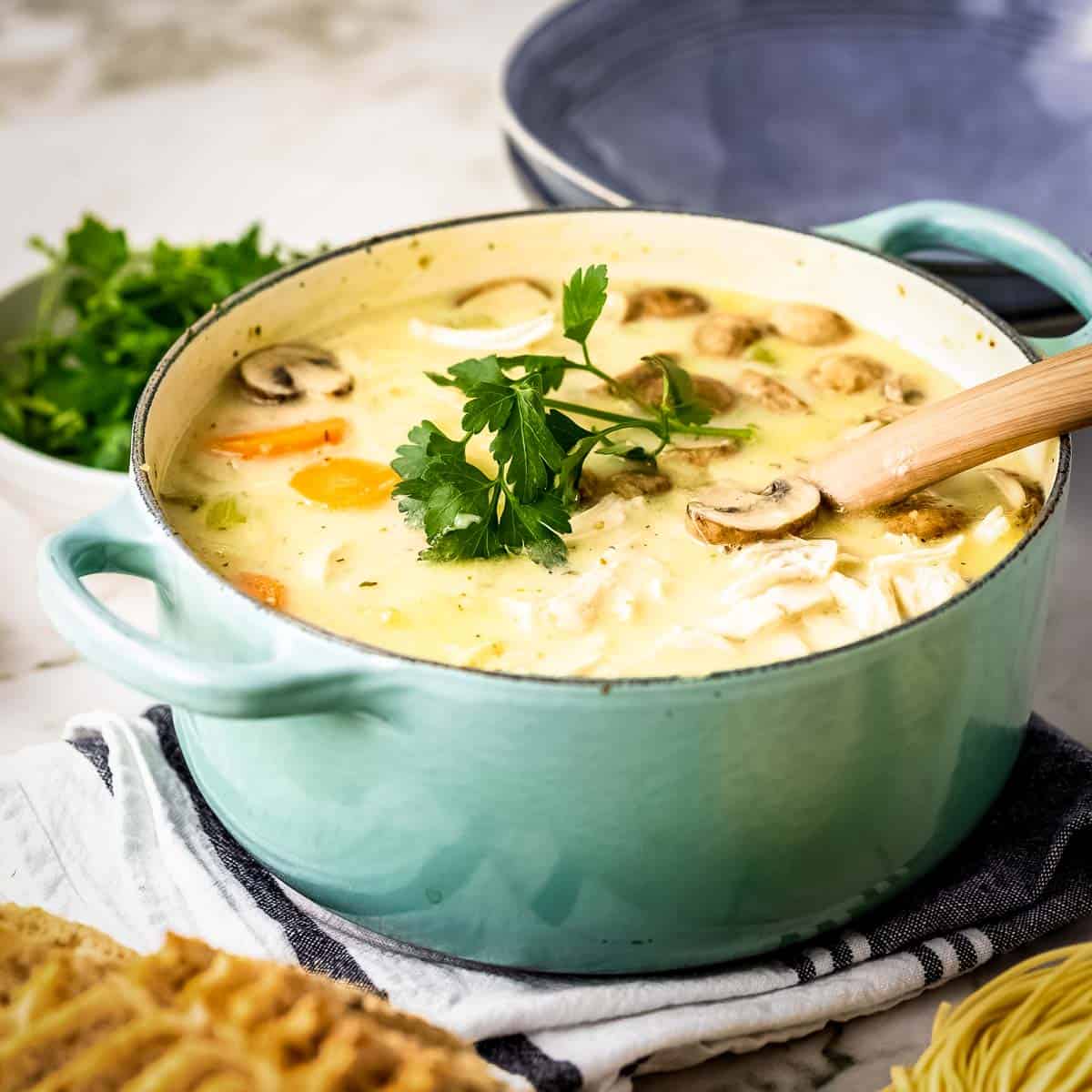 Side shot of a large teal colored pot filled with turkey noodle soup garnished with fresh parsley on a dark blue and white cloth with a wooden spoon leaning on the edge and a bowl of parsley in the background.