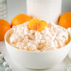 Side shot of orange fluff garnished with mandarin slices in a white bowl with oranges in the background.