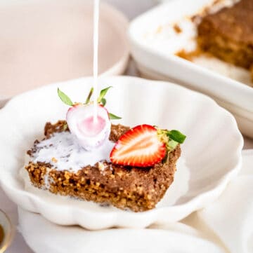 Side close up shot of milk being poured onto quinoa bake garnished with halved strawberries in a white bowl on a white cloth.