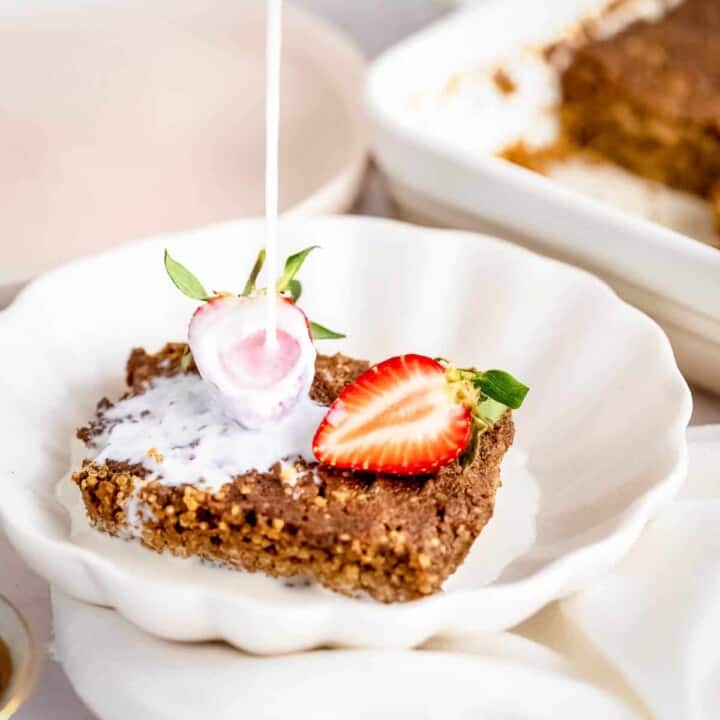 Side close up shot of milk being poured onto quinoa bake garnished with halved strawberries in a white bowl on a white cloth.