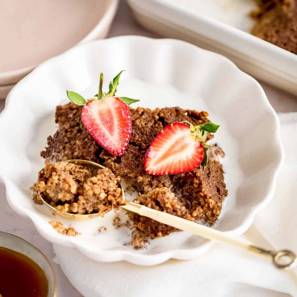 Side close up shot of a gold spoon picking up some quinoa bake out of a white bowl garnished with halved strawberries on a white cloth.