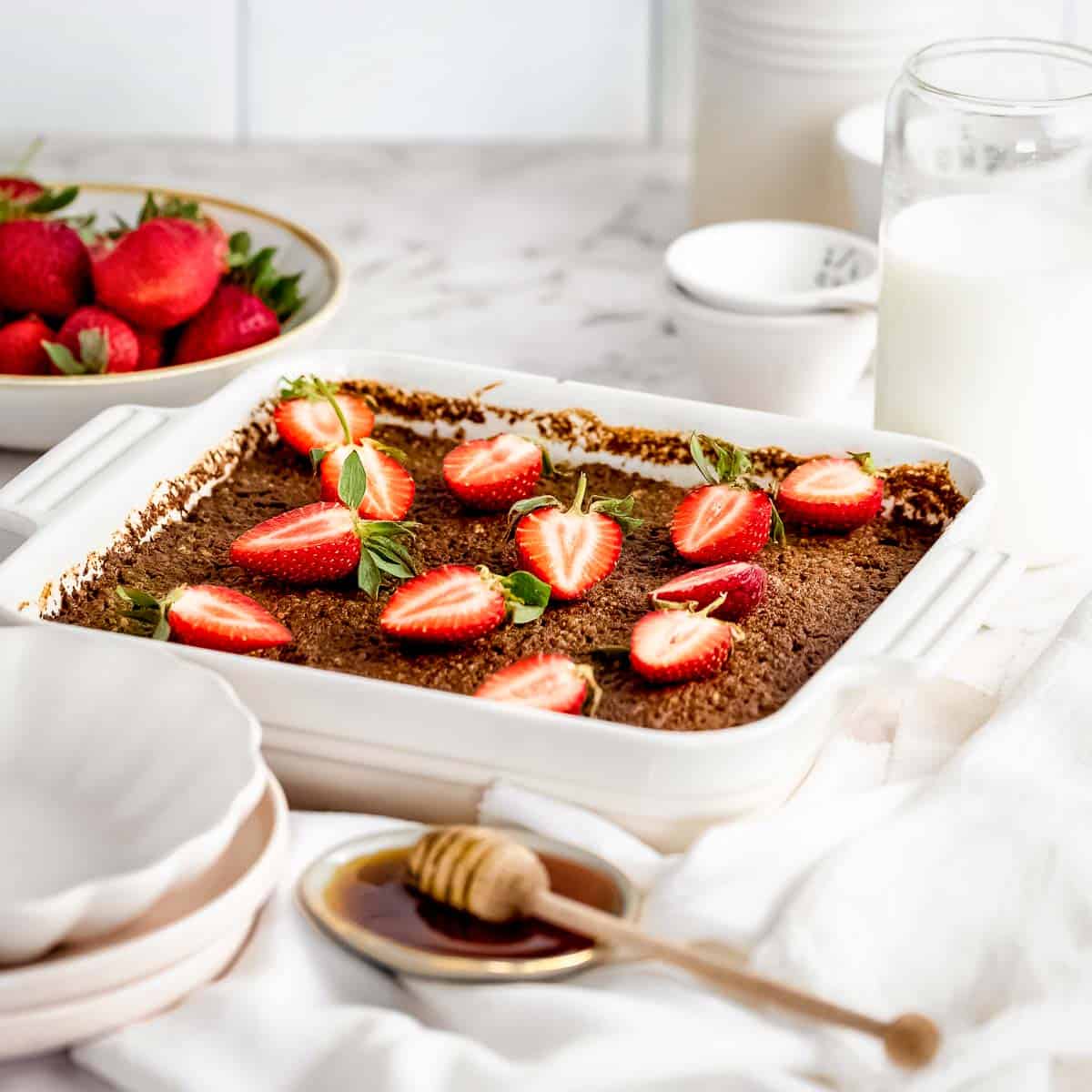 Side shot of a pan of quinoa bake with halved strawberries on top with a shallow bowl with honey on the side with a honey dipper stick all on a white cloth and granite countertop.