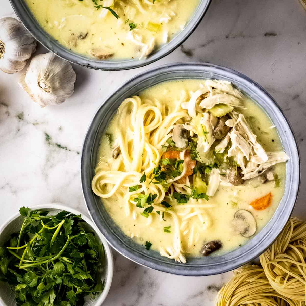 Overhead shot of two grey bowls with turkey noodle soup garnished with fresh herbs on a marble countertop, with a bowl of parsley, garlic cloves, and uncooked egg noodles on the side.