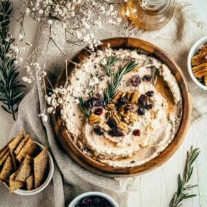 Overhead shot of hummus garnished with pecans, cranberries, and rosemary in a large wooden bowl on a burlap cloth with rosemary, baby breath, and crackers on the side.