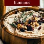 Side shot of hummus garnished with rosemary, pecans, and cranberries in a large wooden bowl on a burlap cloth with crackers, and two jars in the background.