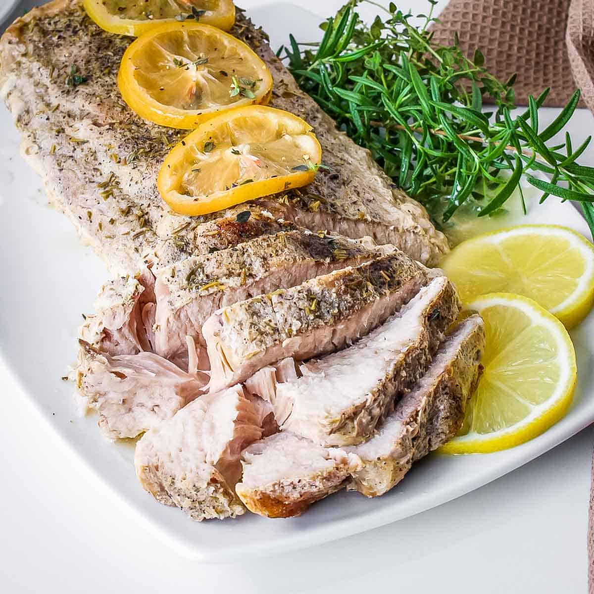 Side shot of a whole pork roast with slices on the side garnished with lemon slices and thyme on a white rectangle plate.