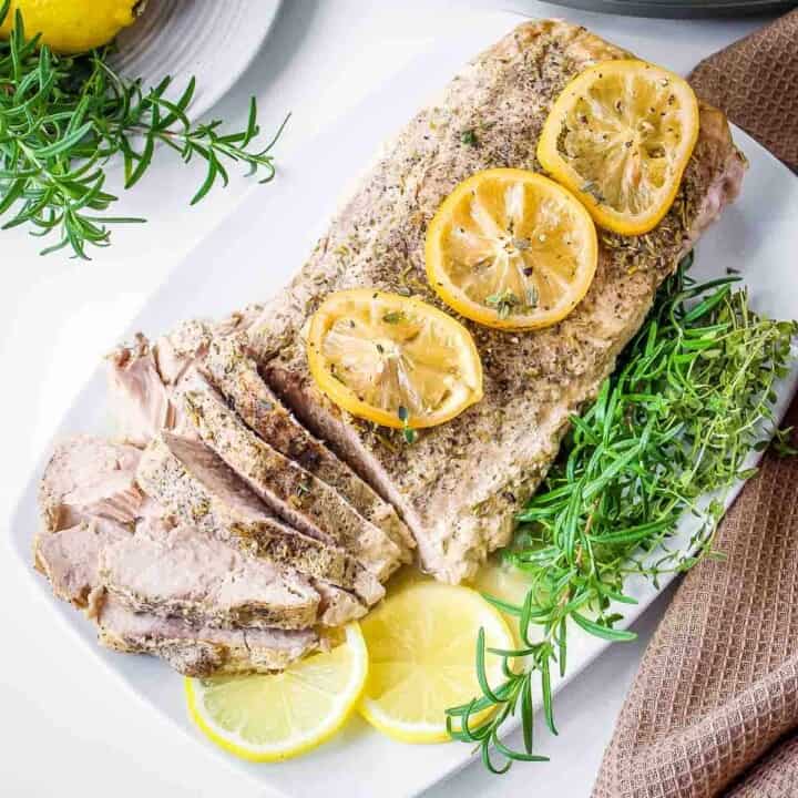 Overhead shot of a whole pork roast with slices on the side garnished with lemon slices and thyme on a white rectangle plate.