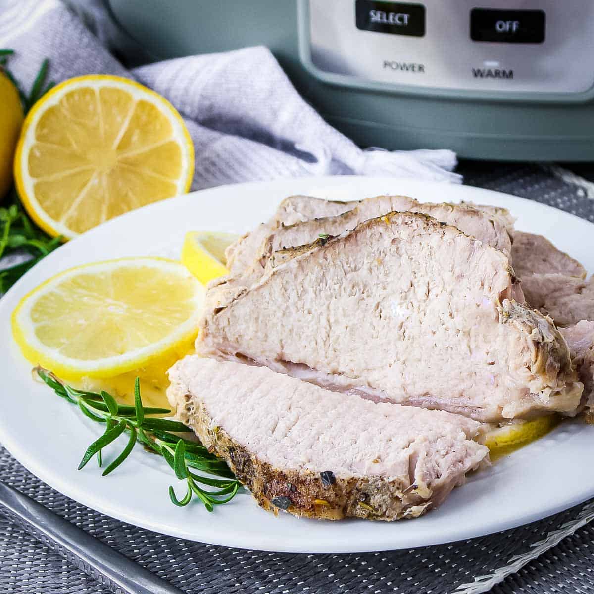 Side shot of slices of pork on a white plate with thyme and lemon slices on the side and the crock pot in the background.