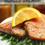 Side shot of two salmon filets garnished with an orange slice on a bed of green beans on a square white plate with a blue cloth and fork on the side.