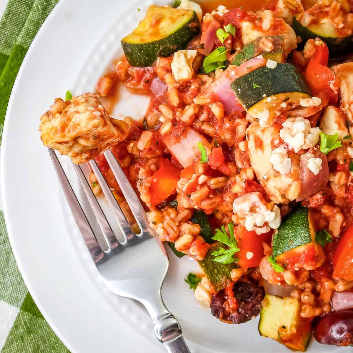 Close up overhead shot of a fork picking up a piece of chicken from a plate of casserole garnished with feta cheese on a white plate.