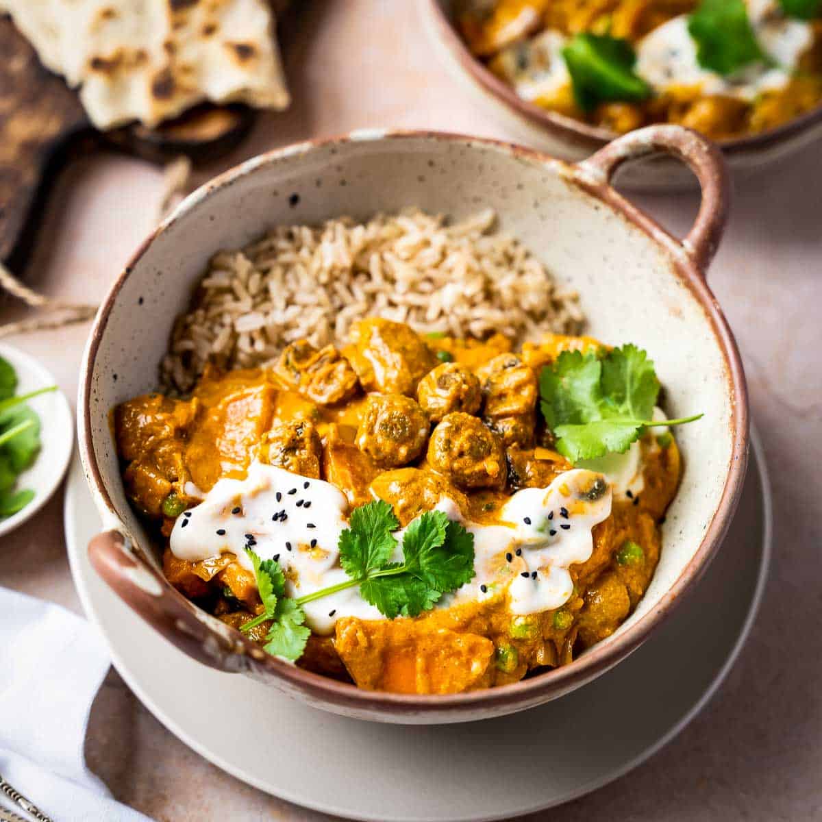 Overhead angled shot of tikka masala with rice in a brown speckled bowl on a grey plate with naan and a wooden cutting board in the background.