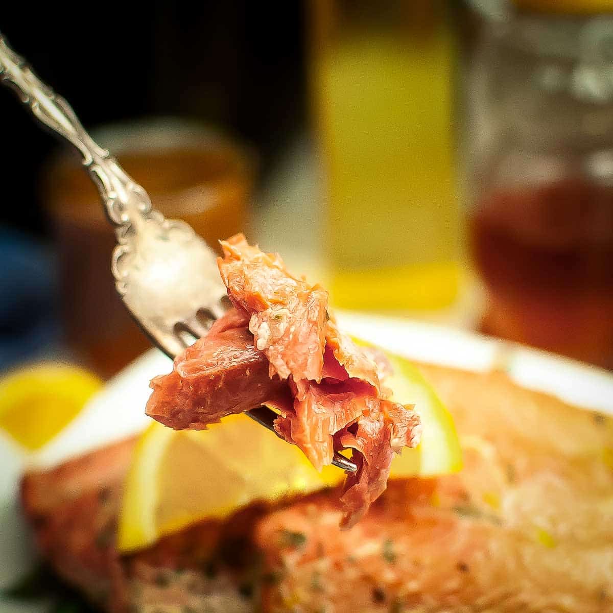 Side close up shot of a fork holding up a piece of salmon over a white plate with two salmon filets garnished with an orange slice.