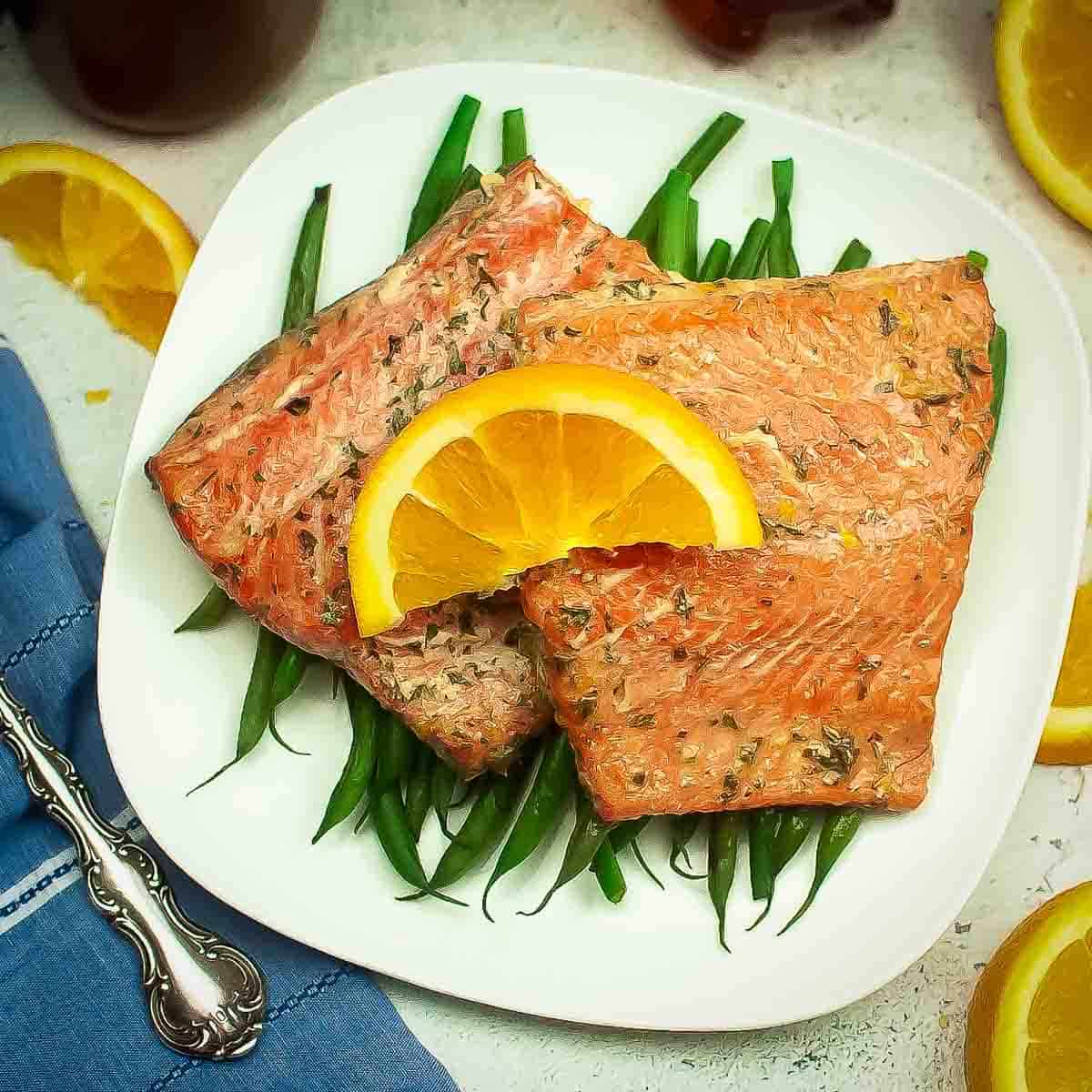 Overhead shot of two salmon filets garnished with an orange on a bed of green beans on a white square plate with a blue cloth and fork on the side.