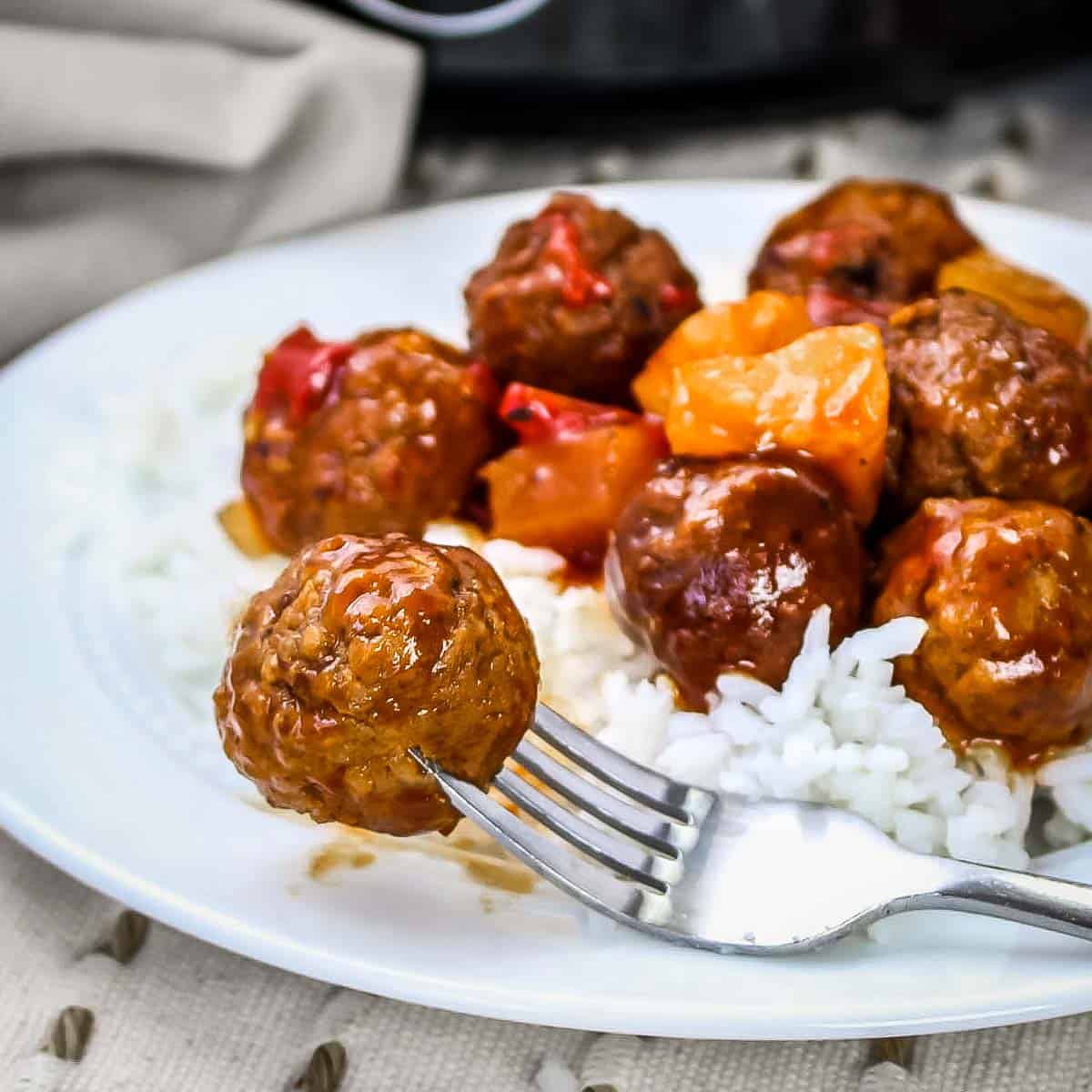Side shot of a fork picking up a meatball on the side of a white plate with white rice and Hawaiian meatballs.