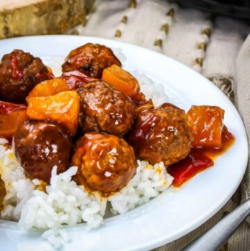 Side angled shot of Hawaiian meatballs over white rice in a white bowl with a white and yellow cloth in the background.