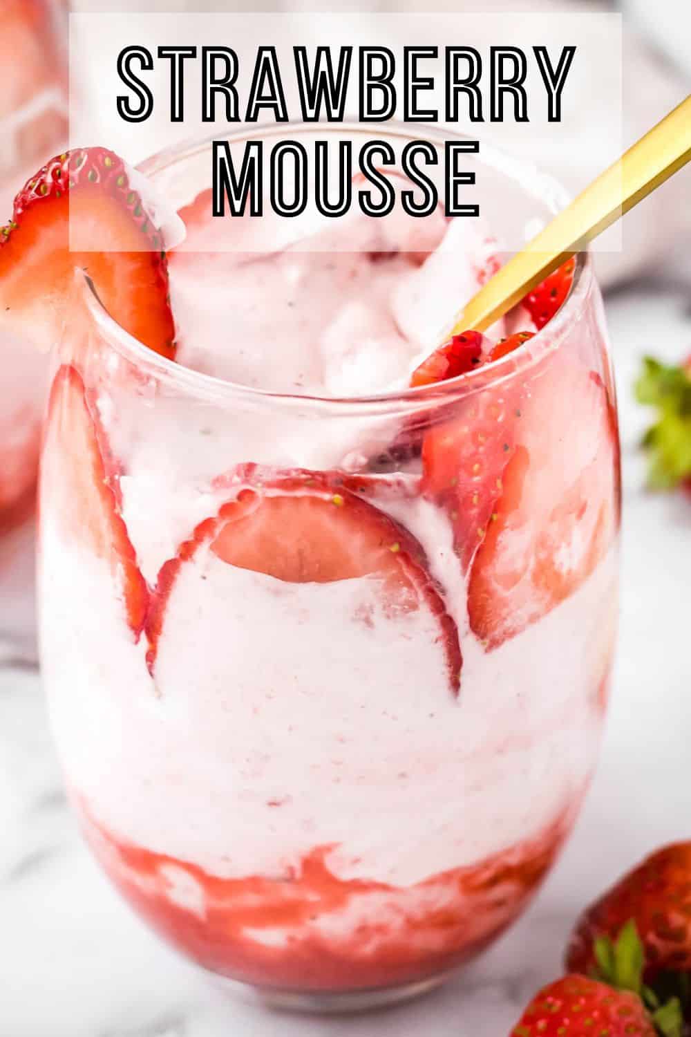 Side close up shot of a glass tumbler with strawberry mousse and sliced strawberries with a gold spoon on a marble table.