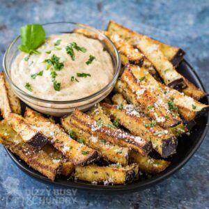 Side angled shot of eggplant fries garnished with basil and parmesan on a black plate with a clear bowl of chipotle basil dip garnished with more fresh basil.
