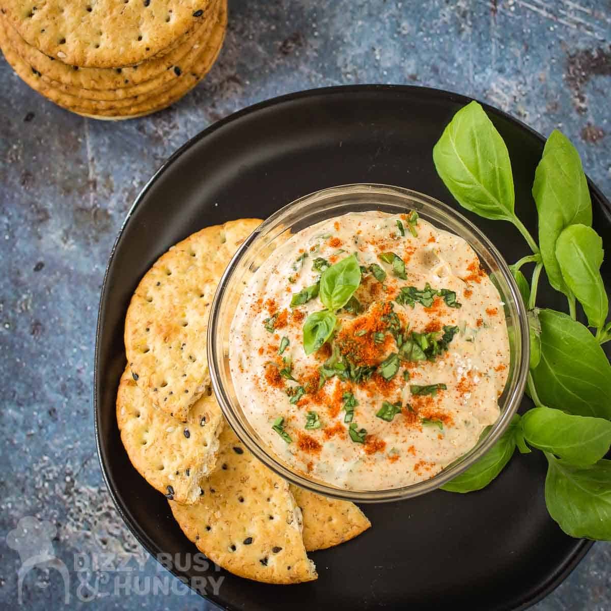Overhead shot of chipotle dip garnished with paprika and basil in a clear bowl on a black plate with halved crackers and basil on the side.