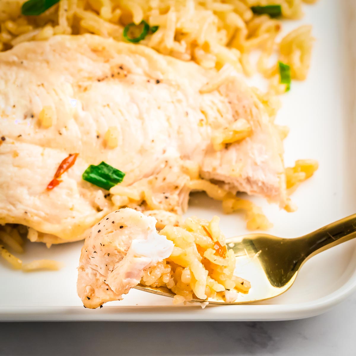 Side shot of a golden fork picking up a piece of chicken and some rice on a white plate with more in the background.